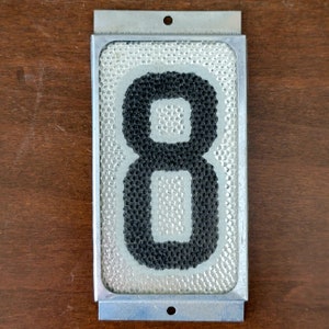 May include: A silver metal house number plaque with a black number eight. The number is made of small black dots on a white background.