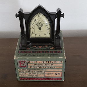 May include: A dark brown New Haven Easel-Artlarm clock with a pointed arch frame and cream-colored face, resting on its original green cardboard box. The box features decorative text including "Easel-Artlarm" and "New Haven Clock Company."