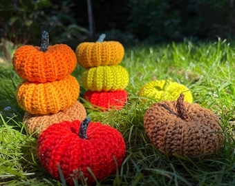 May include: A collection of handmade crochet pumpkins in various fall colors, including orange, yellow, red, and brown. Some are stacked, while others are scattered on green grass, creating a festive autumn display.