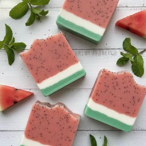 May include: Four rectangular soaps designed to resemble watermelon slices. The soaps feature a pink top with black speckles, a white middle, and a green bottom. Fresh mint leaves and watermelon slices are scattered around the soaps.
