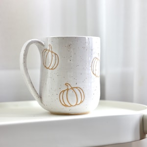 Peut inclure: Une tasse en céramique blanche avec une finition mouchetée et une anse incurvée. La surface extérieure présente plusieurs illustrations de citrouilles en relief de couleur brun clair réparties sur le corps de la tasse.