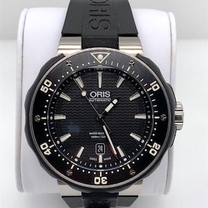 Könnte beinhalten: Eine schwarze Oris Automatic Uhr mit schwarzem Kautschukband und silberner Lünette. Das Zifferblatt der Uhr ist schwarz mit weißen Markierungen und einem Datumsfenster bei 3 Uhr. Die Uhr ist wasserdicht bis 1000 Meter.