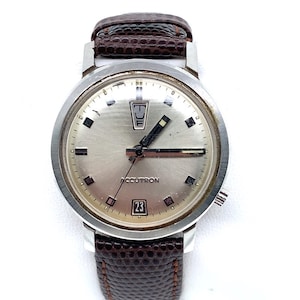 May include: A silver Accutron wristwatch with a white face, black hands, and a brown leather band. The watch face has a day of the week indicator, a date window, and black hour markers.