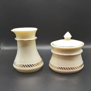 May include: Two cream-colored ceramic containers with gold trim. One is a tall, slender shape with a flared top, and the other is a shorter, rounder shape with a lid. Both have a gold rope-like design around the base.