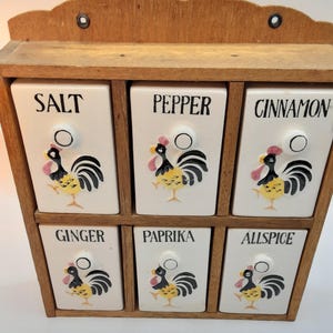 May include: A wooden spice rack with six white ceramic drawers. Each drawer is labeled with a spice name in black text and features a rooster illustration. The rack is made of light brown wood.