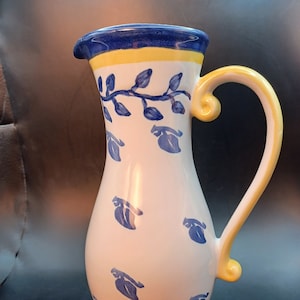 May include: A white ceramic pitcher with a blue and yellow floral design. The pitcher has a wide mouth and a handle.