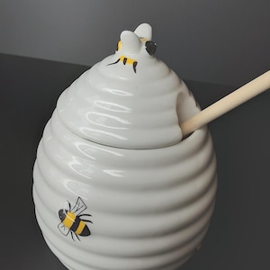 May include: White ceramic honey pot shaped like a beehive, with a wooden honey dipper. The pot has a lid and is decorated with black and yellow bee illustrations. The beehive design has a ridged texture.