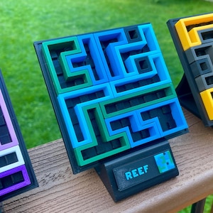 May include: A square 3D printed puzzle with a black base and a maze pattern in blue and green. The base features a black rectangular plaque with the text REEF in blue letters. The puzzle is mounted on a black stand for display.