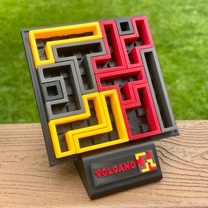 Może przedstawiać: Puzzle wydrukowane w 3D z czarną podstawą i kolorowymi elementami w kolorze ż&oacute;łtym, czerwonym i szarym. Puzzle mają kwadratowy kształt z labiryntowym wzorem. Czarny stojak z napisem "VOLCANO" w kolorze czerwonym z przodu.