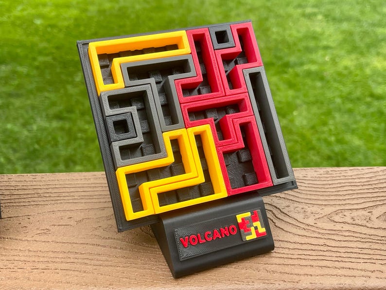 Może przedstawiać: Puzzle wydrukowane w 3D z czarną podstawą i kolorowymi elementami w kolorze ż&oacute;łtym, czerwonym i szarym. Puzzle mają kwadratowy kształt z labiryntowym wzorem. Czarny stojak z napisem "VOLCANO" w kolorze czerwonym z przodu.