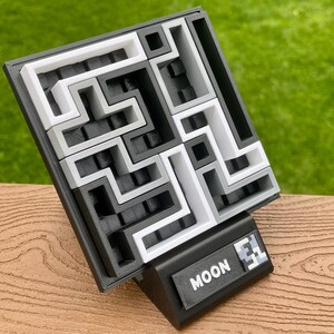 Może przedstawiać: Puzzle 3D z labiryntem w kolorach czarnym, białym i szarym. Puzzle znajduje się na czarnej podstawie z napisem "MOON" i numerem. Tło jest rozmyte zielone.