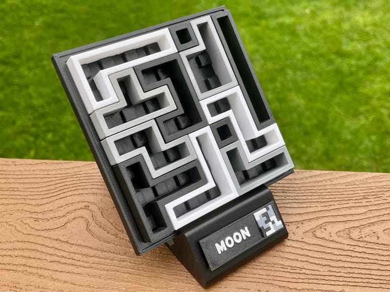 Może przedstawiać: Puzzle 3D z labiryntem w kolorach czarnym, białym i szarym. Puzzle znajduje się na czarnej podstawie z napisem "MOON" i numerem. Tło jest rozmyte zielone.