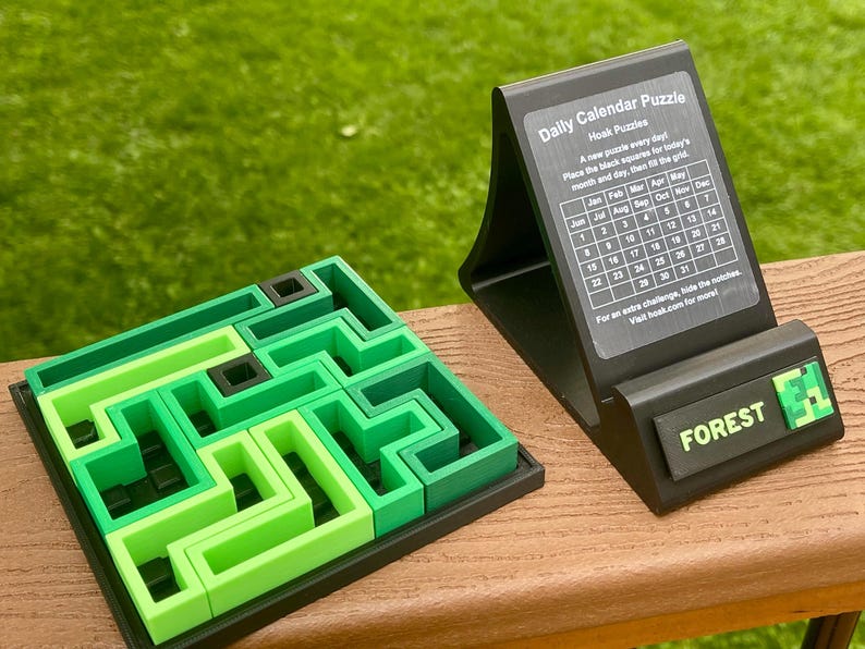 Może przedstawiać: Zielona i czarna układanka z labiryntem obok czarnego stojaka na kalendarz dzienny. Stojak ma siatkę kalendarza i napis "FOREST". Układanka jest wykonana z r&oacute;żnych odcieni zieleni.