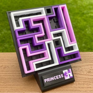 Może przedstawiać: Puzzle 3D z labiryntem w kolorach białym, fioletowym i r&oacute;żowym. Puzzle umieszczone na czarnej podstawie z napisem "PRINCESS" i grafiką. Tło jest rozmyte, zielone.