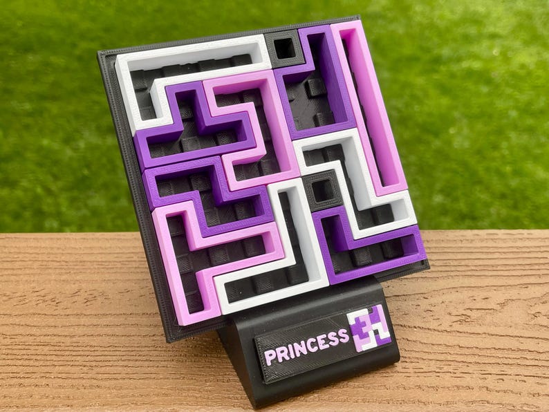 Może przedstawiać: Puzzle 3D z labiryntem w kolorach białym, fioletowym i r&oacute;żowym. Puzzle umieszczone na czarnej podstawie z napisem "PRINCESS" i grafiką. Tło jest rozmyte, zielone.