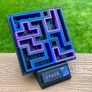 Może przedstawiać: Puzzle wydrukowane w 3D z czarną podstawą i wzorem labiryntu w kolorze niebieskim i fioletowym. Puzzle są podparte czarnym stojakiem z napisem "SPACE". Puzzle mają na celu rzucenie wyzwania użytkownikowi.