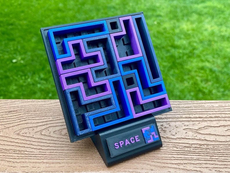 Może przedstawiać: Puzzle wydrukowane w 3D z czarną podstawą i wzorem labiryntu w kolorze niebieskim i fioletowym. Puzzle są podparte czarnym stojakiem z napisem "SPACE". Puzzle mają na celu rzucenie wyzwania użytkownikowi.