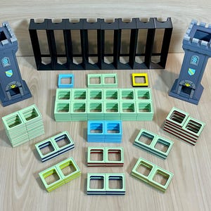 May include: A tabletop game set featuring two gray castle towers, a long black wall section, and numerous square-holed building blocks in light green, blue, yellow, and striped patterns. The components are arranged on a flat surface for assembly.