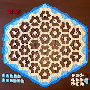 May include: A board game with a hexagonal layout, featuring beige land tiles surrounded by blue water. Includes red, white, and black game pieces, and small blue boat-shaped tokens. The game board is set on a dark brown surface.