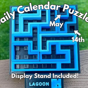 Może przedstawiać: Codzienna układanka kalendarzowa z labiryntem w odcieniach niebieskiego i czarnego. Tekst "Daily Calendar Puzzle!" jest wyświetlany u g&oacute;ry, ze strzałkami wskazującymi "May 14th". Podstawa wyświetlacza na dole zawiera napis "Display Stand Included!" i "LAGOON".