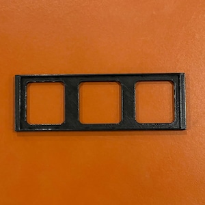 May include: A black plastic frame with three rectangular openings. The frame is designed to hold three small items, such as photos or cards.
