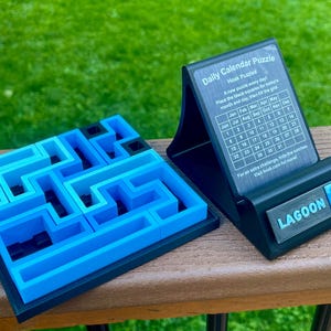Może przedstawiać: Niebiesko-czarna układanka labiryntu i czarna codzienna układanka kalendarza. Kalendarz ma napis "Daily Calendar Puzzle" i "LAGOON" na podstawie. Układanka labiryntu jest kwadratowa.