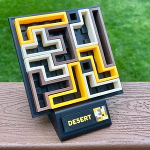 Może przedstawiać: Puzzle 3D z czarną podstawą i wzorem labiryntu w kolorze ż&oacute;łtym, brązowym i ecru. Puzzle zatytułowane "DESERT" w ż&oacute;łtym tekście na czarnej podstawie.