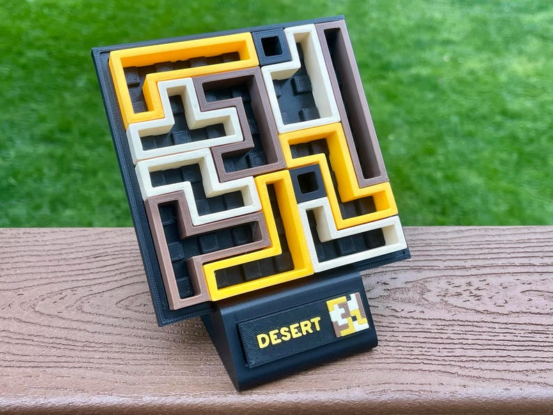 Może przedstawiać: Puzzle 3D z czarną podstawą i wzorem labiryntu w kolorze ż&oacute;łtym, brązowym i ecru. Puzzle zatytułowane "DESERT" w ż&oacute;łtym tekście na czarnej podstawie.