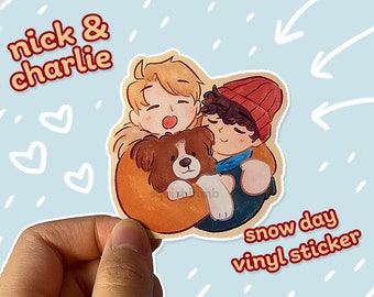 Op de afbeelding: Een vinyl sticker met een cartoonillustratie van twee mensen en een hond. De mensen knuffelen elkaar en de hond. De mensen dragen winterkleding en de hond is bruin en wit. De sticker is op een blauwe achtergrond met witte regendruppels en de tekst "nick & charlie" en "snow day vinyl sticker".