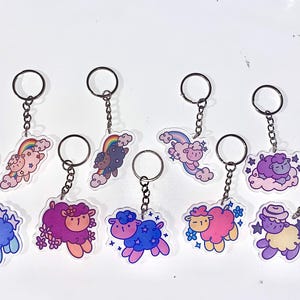 May include: A set of ten colourful keychains featuring cartoon sheep in various poses. Each keychain has a silver metal ring and a clear acrylic charm. The sheep are depicted on a cloud, a rainbow, or with stars.
