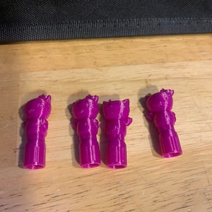 May include: Four small, pink, 3D printed figurines of a cartoon character. The figurines are standing upright on a wooden surface.