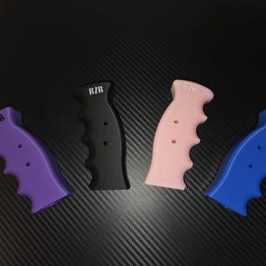 May include: Four contoured gear shifter handles in purple, black, light pink, and blue. Each handle features a finger-grip design, two small circular holes, and the text RZR printed in white near the top of the grip surface.