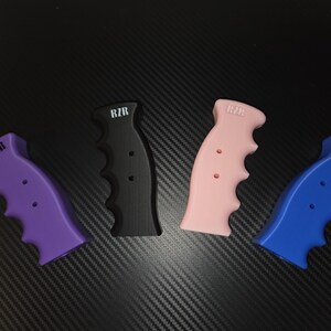 May include: Four contoured gear shifter handles in purple, black, light pink, and blue. Each handle features a finger-grip design, two small circular holes, and the text RZR printed in white near the top of the grip surface.