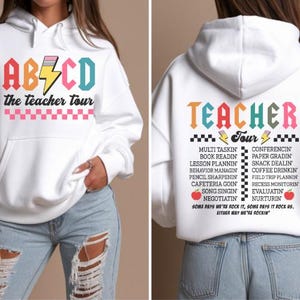 May include: A white hooded sweatshirt featuring a front graphic with the text ABCD, a pencil icon, and the teacher tour. The back displays the text TEACHER TOUR, a list of tasks, and the phrase SOME DAYS WE'RE ROCK IT, SOME DAYS IT ROCK US.