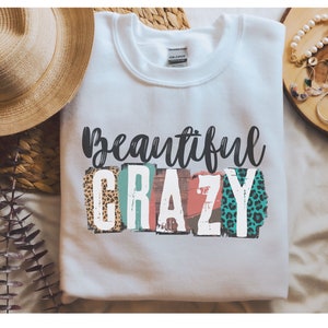 May include: White sweatshirt with the text "Beautiful Crazy" in black lettering. The letters are decorated with colorful, abstract patterns, including leopard print.