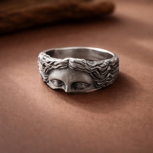 Può includere: Un anello in metallo color argento con un rilievo scolpito di un volto umano con capelli dettagliati e fluenti. La fascia presenta una finitura ossidata che mette in risalto i tratti del viso, inclusi gli occhi, il naso e la texture.