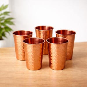 Puede incluir: Cinco vasos de color cobre con una textura martillada. Los vasos son cilíndricos con un borde ligeramente ensanchado. Están dispuestos sobre una superficie de madera de color claro.
