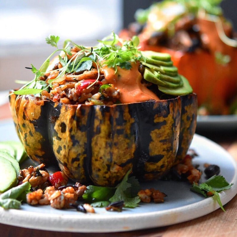 May include: A close-up of a stuffed acorn squash dish. The squash is dark green and yellow with a filling of rice, vegetables, and a creamy sauce. Sliced avocado and fresh herbs garnish the dish, which is served on a light blue plate.