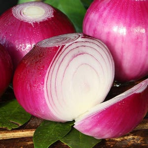 May include: Close-up of several red onions, one cut in half to reveal concentric white and red rings. The onions have a vibrant pinkish-red skin and are surrounded by green leaves, suggesting a fresh produce theme.