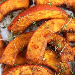 May include: Close-up of roasted pumpkin wedges, showcasing vibrant orange flesh and a slightly charred exterior. Sprigs of fresh green herbs are scattered across the pieces. A delicious and healthy food.
