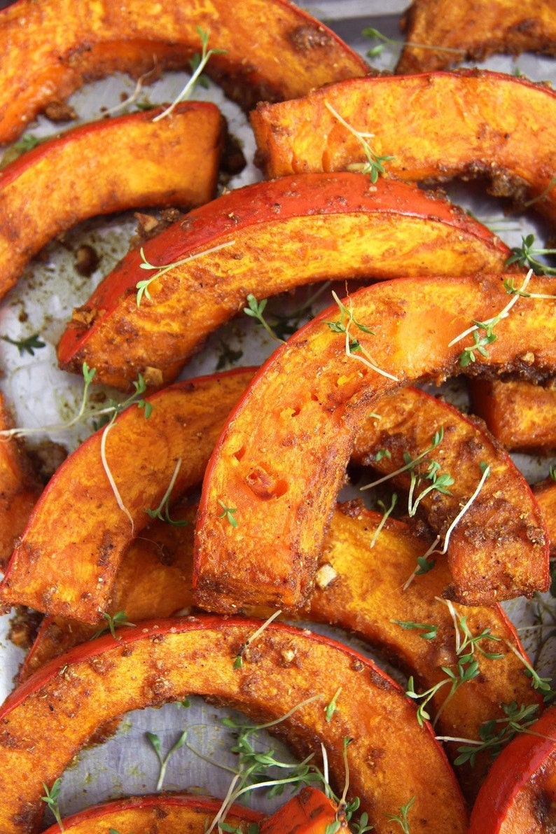 May include: Close-up of roasted pumpkin wedges, showcasing vibrant orange flesh and a slightly charred exterior. Sprigs of fresh green herbs are scattered across the pieces. A delicious and healthy food.