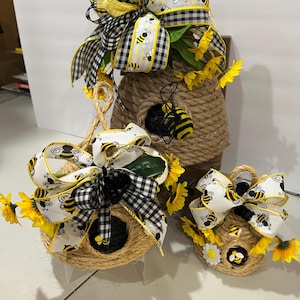 May include: Three decorative bee-themed items. A large beehive-shaped structure and two smaller birdhouse-shaped items are wrapped in rope. Each has a black and white checkered bow with yellow accents and bee details, along with artificial sunflowers.