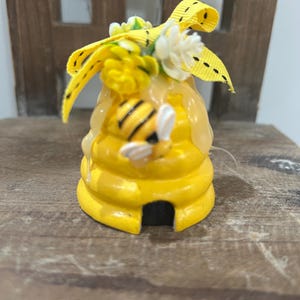 May include: A yellow, beehive-shaped ornament with a small opening at the base. The ornament is decorated with a yellow and black striped bee, yellow flowers, and a yellow ribbon. The ornament is sitting on a wooden surface.