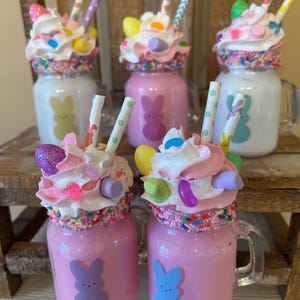 May include: Five mason jars filled with pink and white frosting, candy, and sprinkles. Each jar has a different colored bunny sticker on the front. The jars are arranged in a row on a wooden surface.
