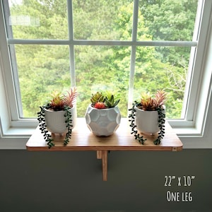 May include: A wooden shelf with a single leg, measuring 56 cm by 25 cm, holds three white ceramic planters with succulent plants. The shelf is mounted on a windowsill with a view of green trees.