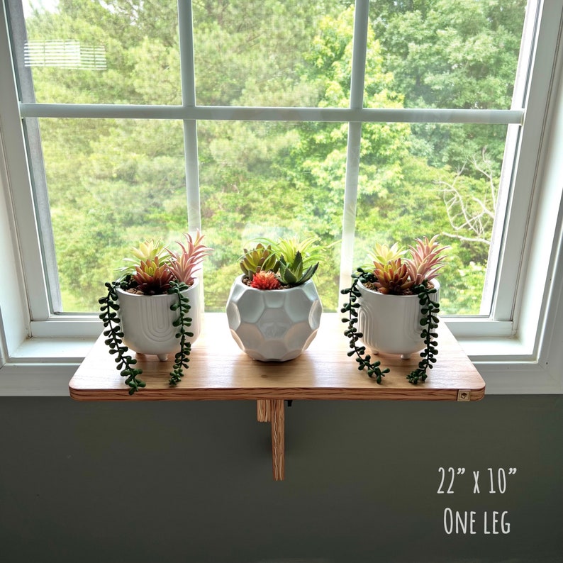 May include: A wooden shelf with a single leg, measuring 56 cm by 25 cm, holds three white ceramic planters with succulent plants. The shelf is mounted on a windowsill with a view of green trees.