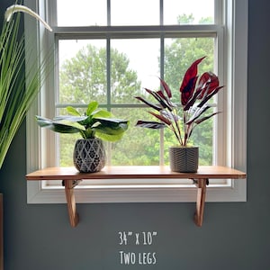 May include: A wooden window shelf with two legs, measuring 86 cm by 25 cm. The shelf is mounted below a window and holds two potted plants. The shelf is made of natural wood and has a light brown finish.