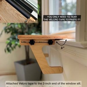 K&ouml;nnte beinhalten: Ein Holzregal mit verstellbaren Halterungen und schwarzen Kn&ouml;pfen, das mit Klettband an einer Fensterbank befestigt ist. Das Regal ist so konzipiert, dass es an einer Fensterbank montiert werden kann, wobei die Halterungen nach au&szlig;en ragen, um das Regal zu st&uuml;tzen. Das Regal ist aus Naturholz gefertigt und hat ein schlichtes, modernes Design. Der Text "YOU ONLY NEED TO TEAR THE VELCRO TAPE TO PASTE IT!" ist auf dem Bild zu sehen.