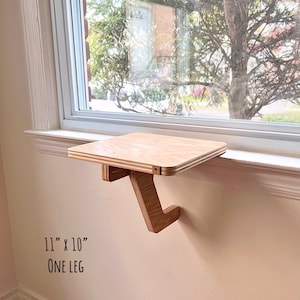 May include: A wooden shelf with a single leg, measuring 11 inches by 10 inches, mounted on a windowsill. The shelf is made of light wood and has a natural finish. The text "ONE LEG" is printed below the shelf.