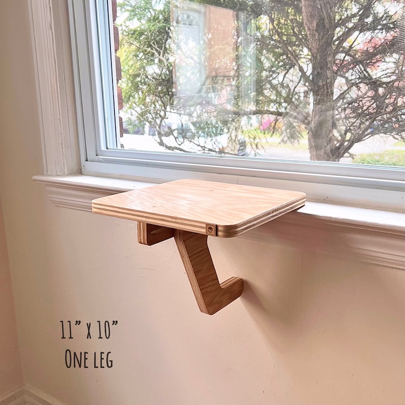 May include: A wooden shelf with a single leg, measuring 11 inches by 10 inches, mounted on a windowsill. The shelf is made of light wood and has a natural finish. The text "ONE LEG" is printed below the shelf.